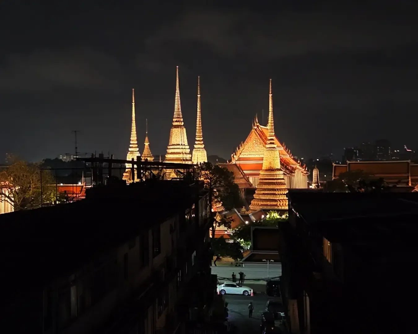 Illuminated Wat Pho temple spires and buildings at night in Bangkok, with a darkened foreground and city skyline in the background. Peaceful ambiance.