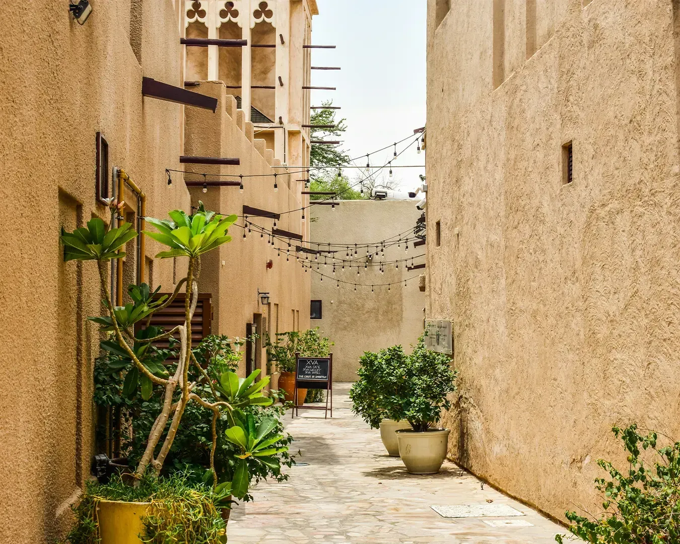 Narrow alley with sandy-colored walls, potted plants, and string lights overhead. Warm, inviting atmosphere with a chalkboard sign in the distance.
