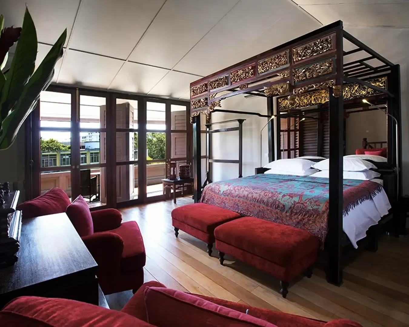 Luxurious bedroom with a carved four-poster bed, elegant red upholstery, and wood furnishings. Sunlit balcony visible through large windows. Cozy and refined.