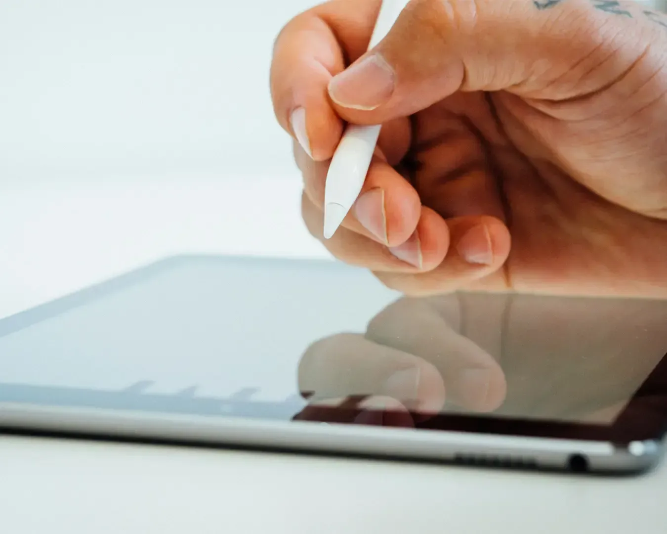 A hand holding a stylus pen hovers over a tablet screen, suggesting digital drawing or note-taking. The setting is minimal and focused on creativity.