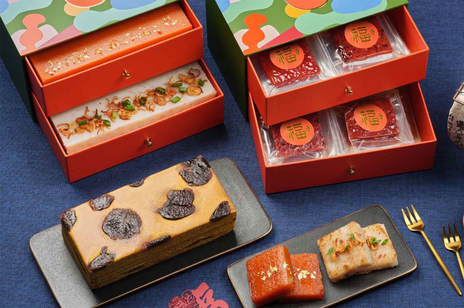 A festive Chinese New Year food flat lay featuring a tiered gift box containing Bak Kwa (dried meat) and Nian Gao, accompanied by a loaf of Kueh Lapis layer cake and fried radish cake.