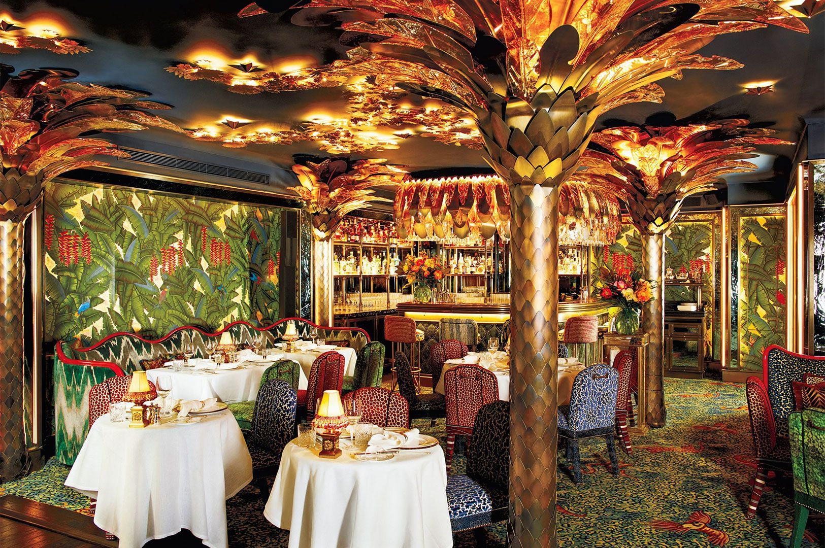An opulent, maximalist restaurant interior with a dark ceiling, featuring gold palm tree columns, leopard-print and patterned chairs, and a bar area in the background.