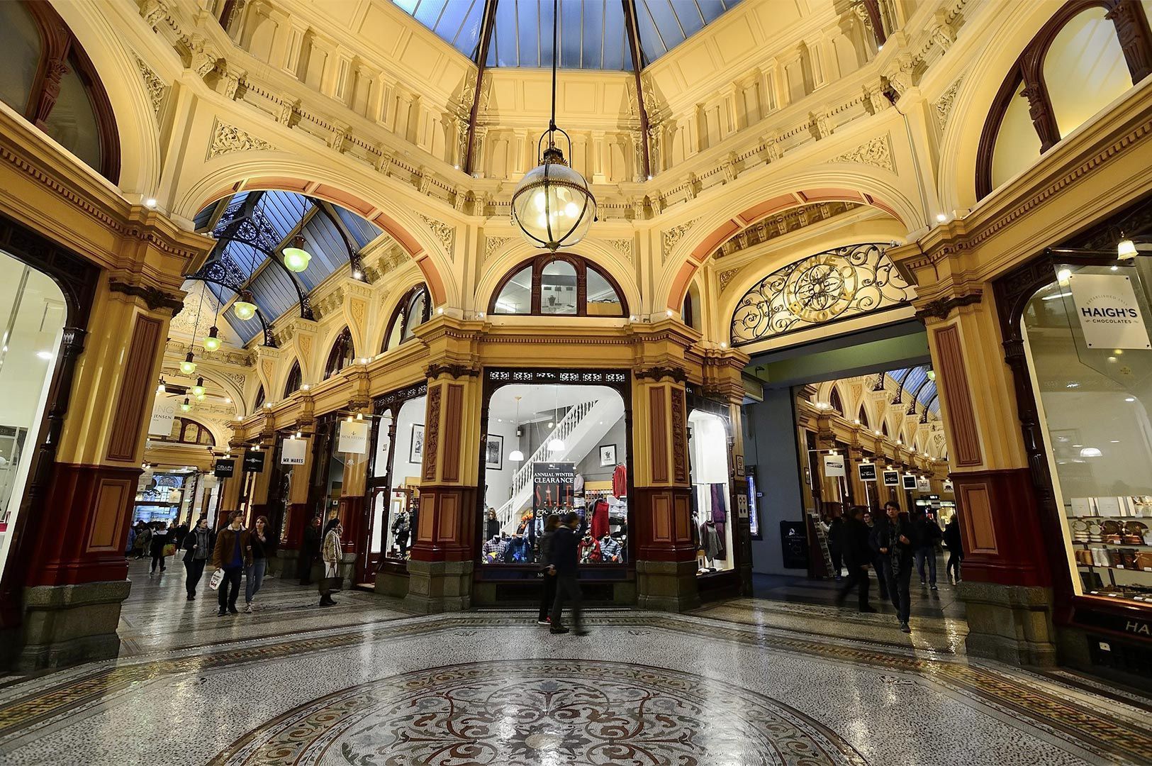 Grand interior of a historic Victorian shopping arcade featuring ornate yellow arches, a glass skylight, and detailed mosaic flooring.