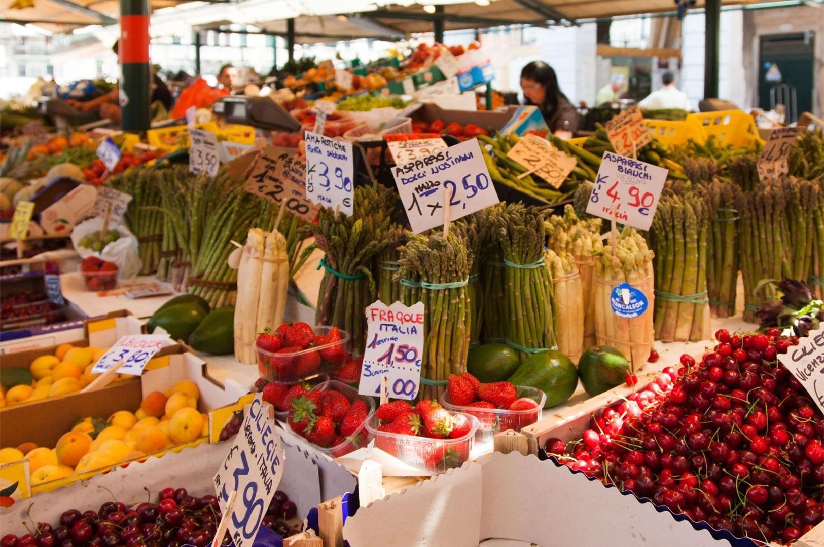 A vibrant display of fresh Italian produce at a market stall, featuring strawberries, cherries, asparagus bundles, lemons, and price tags in Euros.