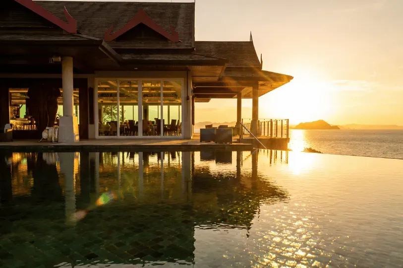 A tranquil infinity pool at sunset perfectly reflecting the golden sky and the adjacent Thai-style luxury villa, which features floor-to-ceiling windows revealing a dining room.
