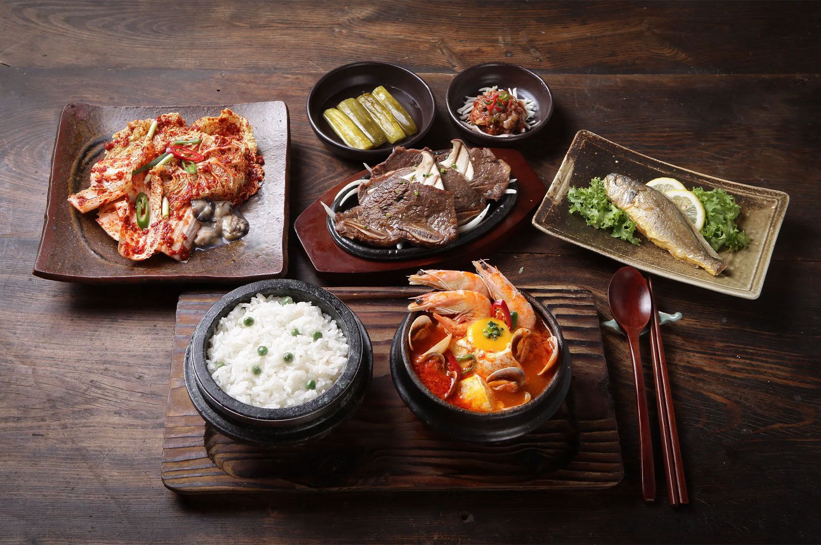 An overhead spread of a complete Korean meal set on a dark wooden board, featuring a bowl of steaming rice, a spicy seafood soft tofu stew (Soon-dub-u Jjigae), grilled Galbi short ribs, kimchi, and a fried fish.