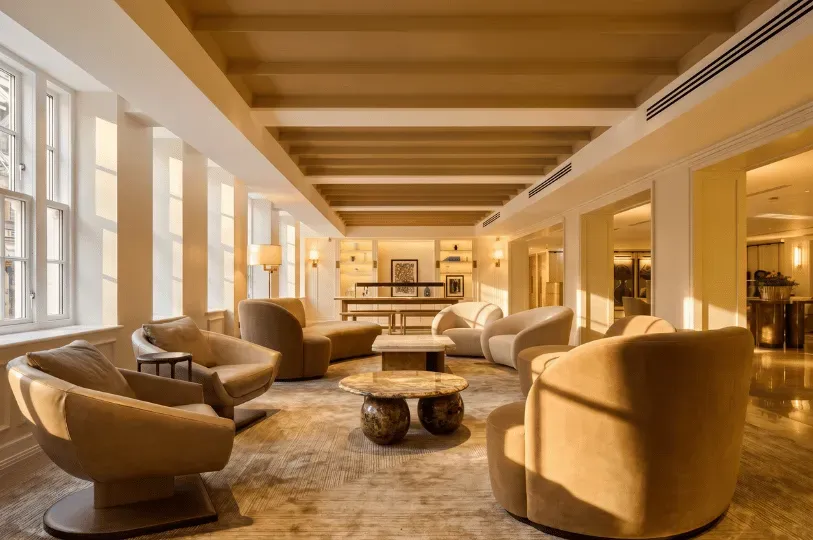 A serene, minimalist lounge washed in golden afternoon light, featuring soft, rounded beige leather armchairs arranged on a large textured rug. Sunlight streams through a row of classic white-framed windows, highlighting the clean lines of the wooden slatted ceiling.