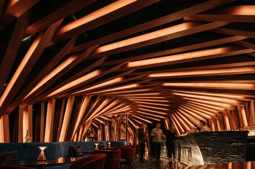 The interior of a moody, dimly lit Melbourne bar featuring a dramatic, cave-like ceiling made of angled wooden beams and warm ambient lighting.