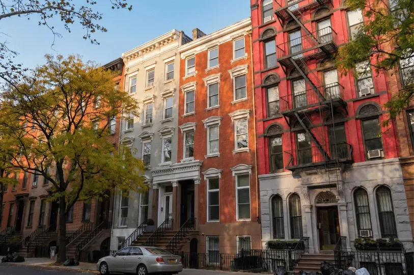 A street-level view of a row of historic walk-up apartment buildings in varied brick colors, featuring classic New York architecture like stoops, fire escapes, and tree-lined sidewalks.