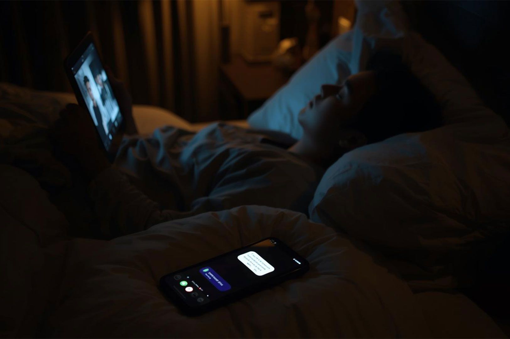 A person lying in bed at night using a tablet, while a smartphone rests on the duvet in the foreground displaying a bright message notification screen.