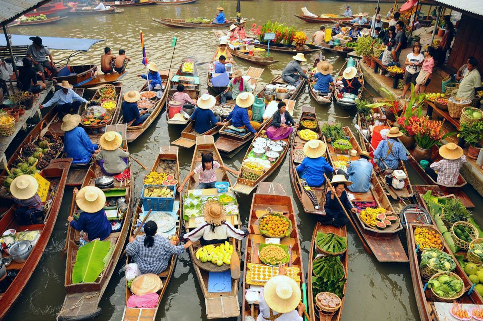 An aerial view of a bustling floating market filled with wooden boats, where vendors in straw hats sell colorful fresh fruits, vegetables, and food on a canal.