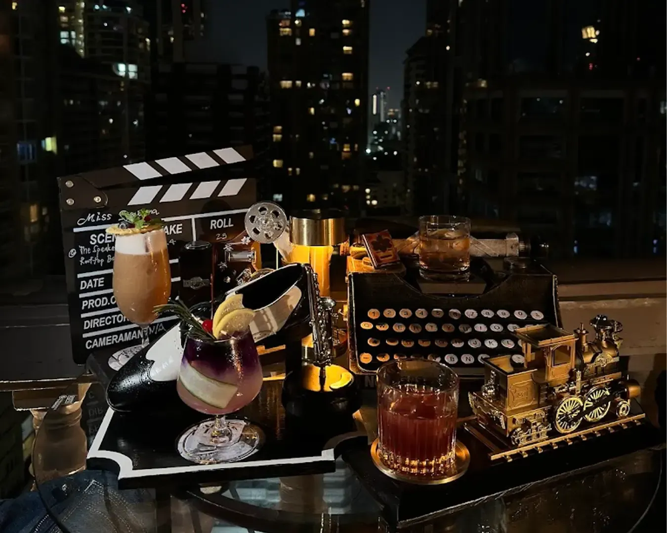 A colorful array of cocktails on a rooftop patio at night, accompanied by a vintage typewriter, film reel, clapperboard, and model train.