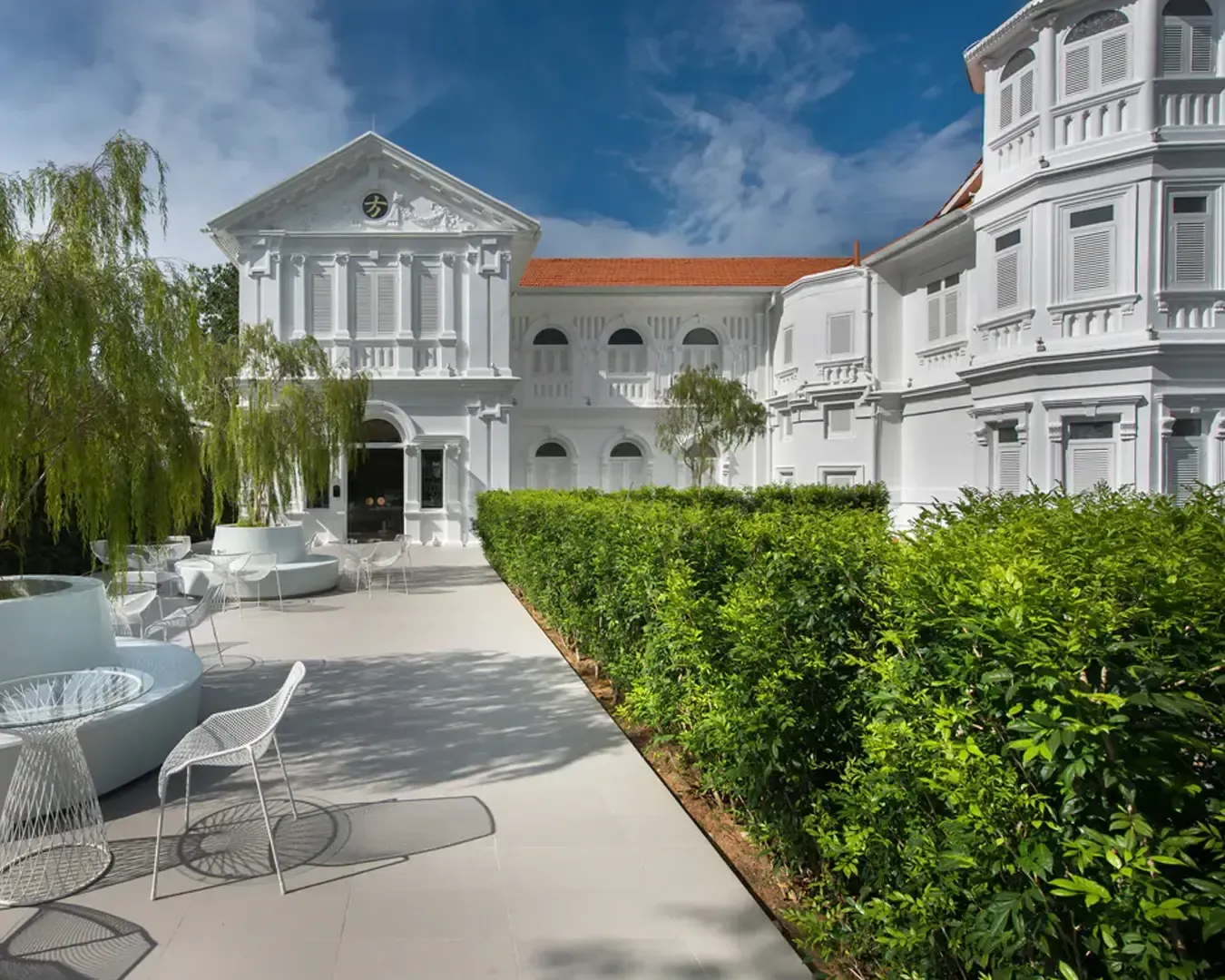 A grand, white colonial building with red tile roof, surrounded by lush greenery and modern outdoor seating under a bright blue sky, conveying elegance.