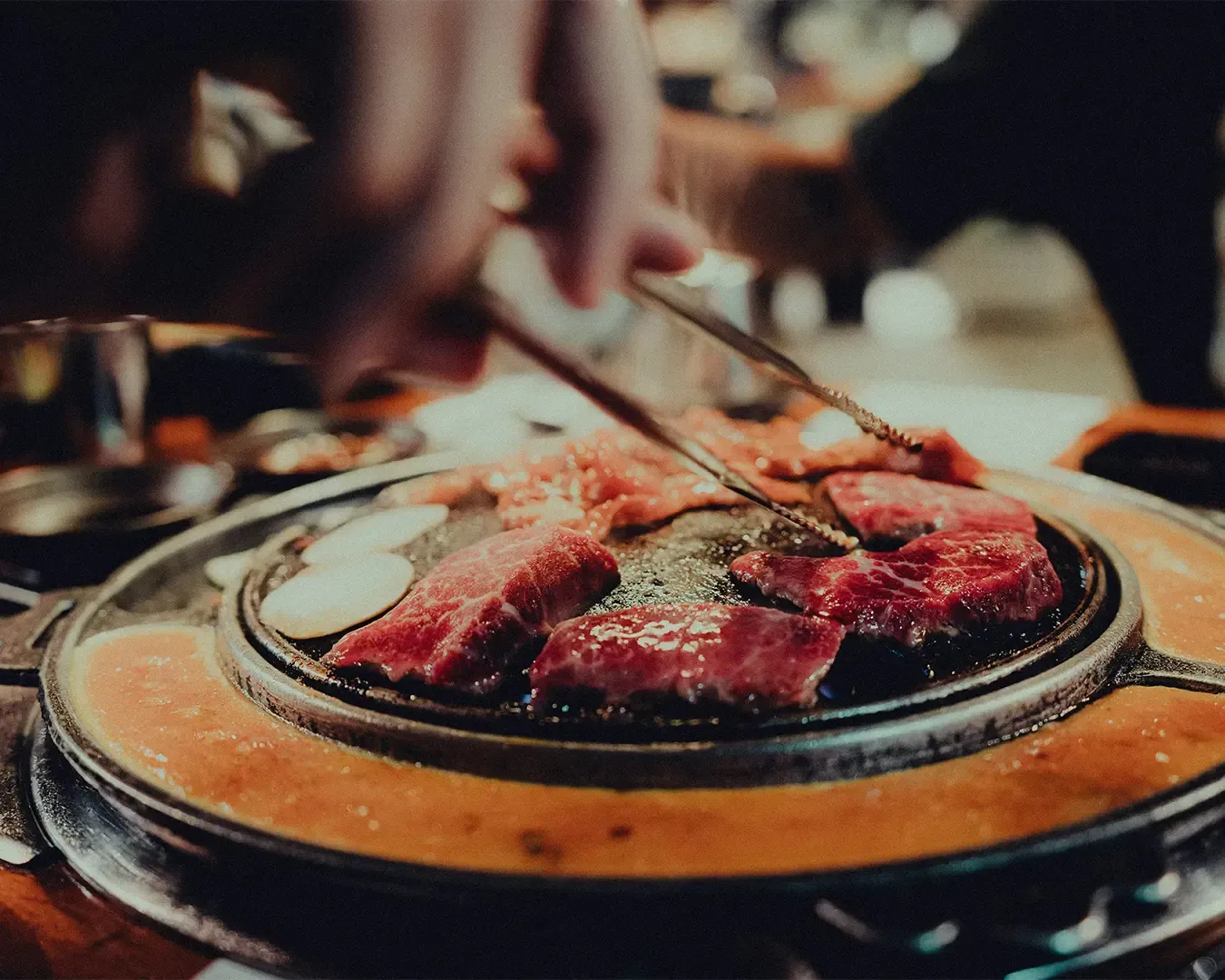 Close-up of sizzling meat grilling over a Korean BBQ, with smoke rising and juices caramelising on the surface. The setting suggests a communal dining experience, with side dishes and diners gathered around the table.