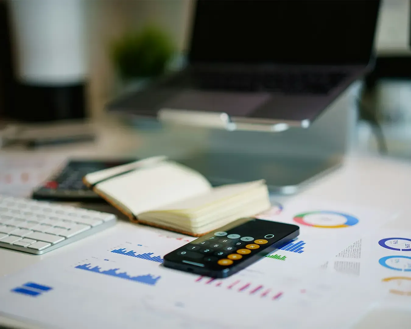 A desk with a laptop, open notebook, smartphone calculator, charts, and keyboard, conveying a business or financial analysis setting.