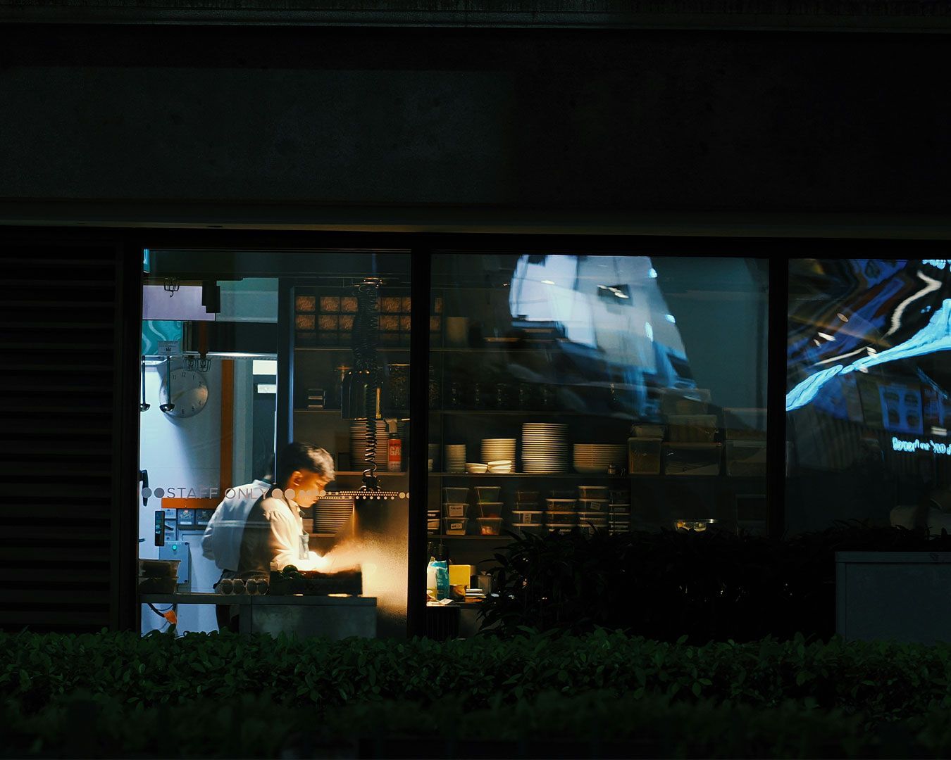 A chef working in a quiet kitchen at night, focused on preparing food under dim overhead lights with cooking tools and pots surrounding the workspace.