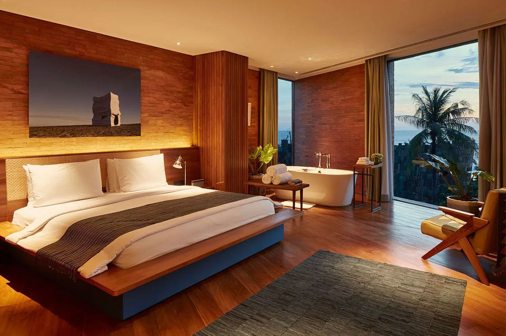 A warm, modern coastal hotel room with exposed red brick walls, a wooden floor, a king-size bed, and a standalone bathtub next to a large window overlooking the ocean and palm trees.