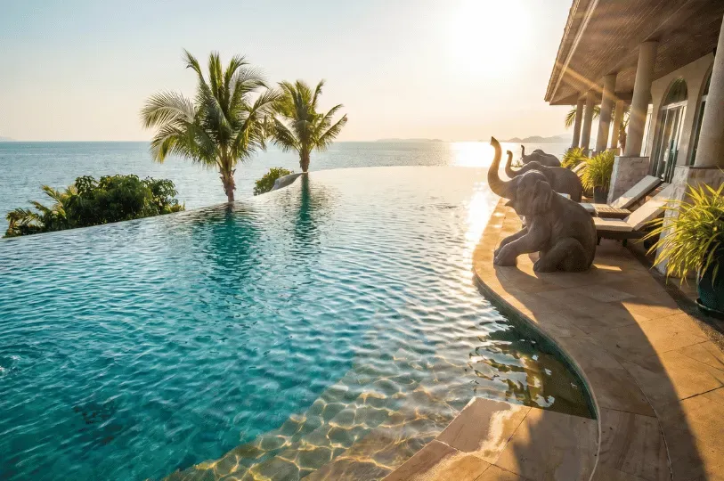 Three sculpted stone elephant statues sitting along the curved edge of an infinity pool overlooking the ocean, set against a stunning tropical sunset and palm trees.
