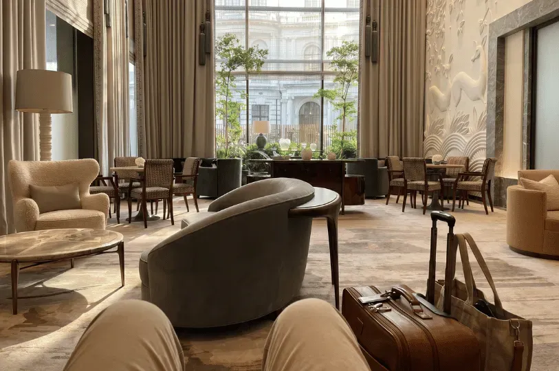 A first-person perspective from a guest sitting in a high-ceilinged hotel lobby lounge, with a leather suitcase and tote bag in the foreground. The room features elegant velvet armchairs, large neutral drapery, a relief wall sculpture of deer, and massive windows overlooking historic London architecture.
