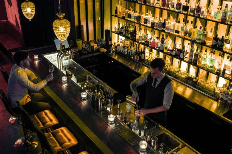 A bartender shaking a cocktail in an atmospheric, speakeasy-style bar with glowing, backlit liquor shelves and ornate vintage pendant lights.