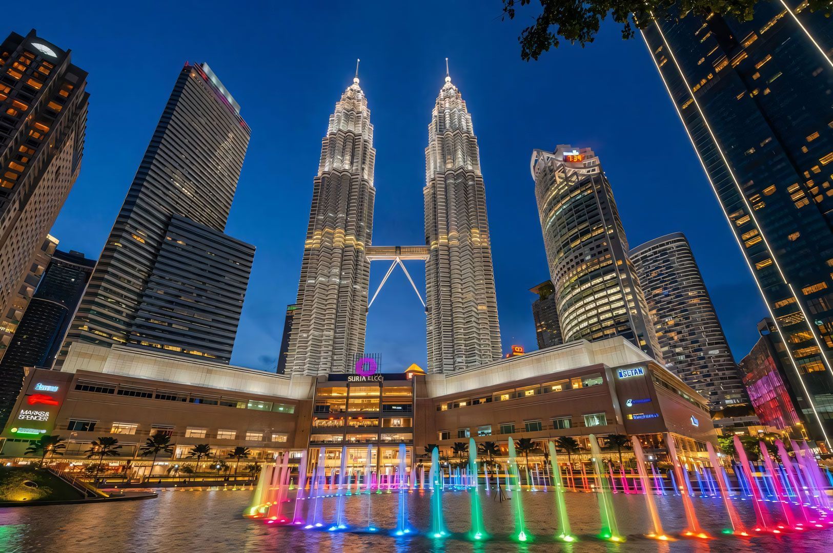 A wide-angle night shot of the majestic Petronas Twin Towers in Kuala Lumpur, rising above the Suria KLCC mall and the colorful Lake Symphony water fountain display.