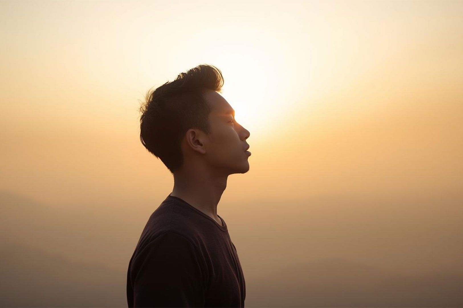 Side profile of a young man, silhouetted or strongly backlit by a bright, golden-orange sunset or sunrise sky, appearing contemplative or peaceful.