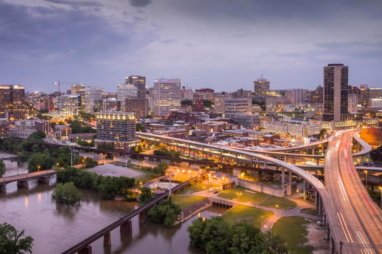 The Richmond, Virginia city skyline at dusk with the James River, bridges, and glowing highway traffic lights.