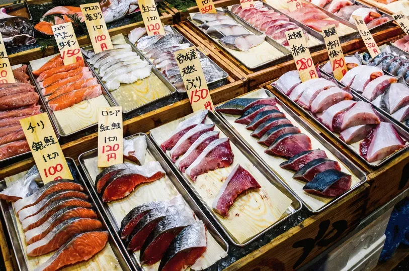 A vibrant fish market display featuring rows of exceptionally fresh, clean-cut fish fillets—including salmon, tuna, and mackerel—arranged neatly on ice trays with handwritten Japanese price tags, representing the premium sourcing behind high-end sushi.