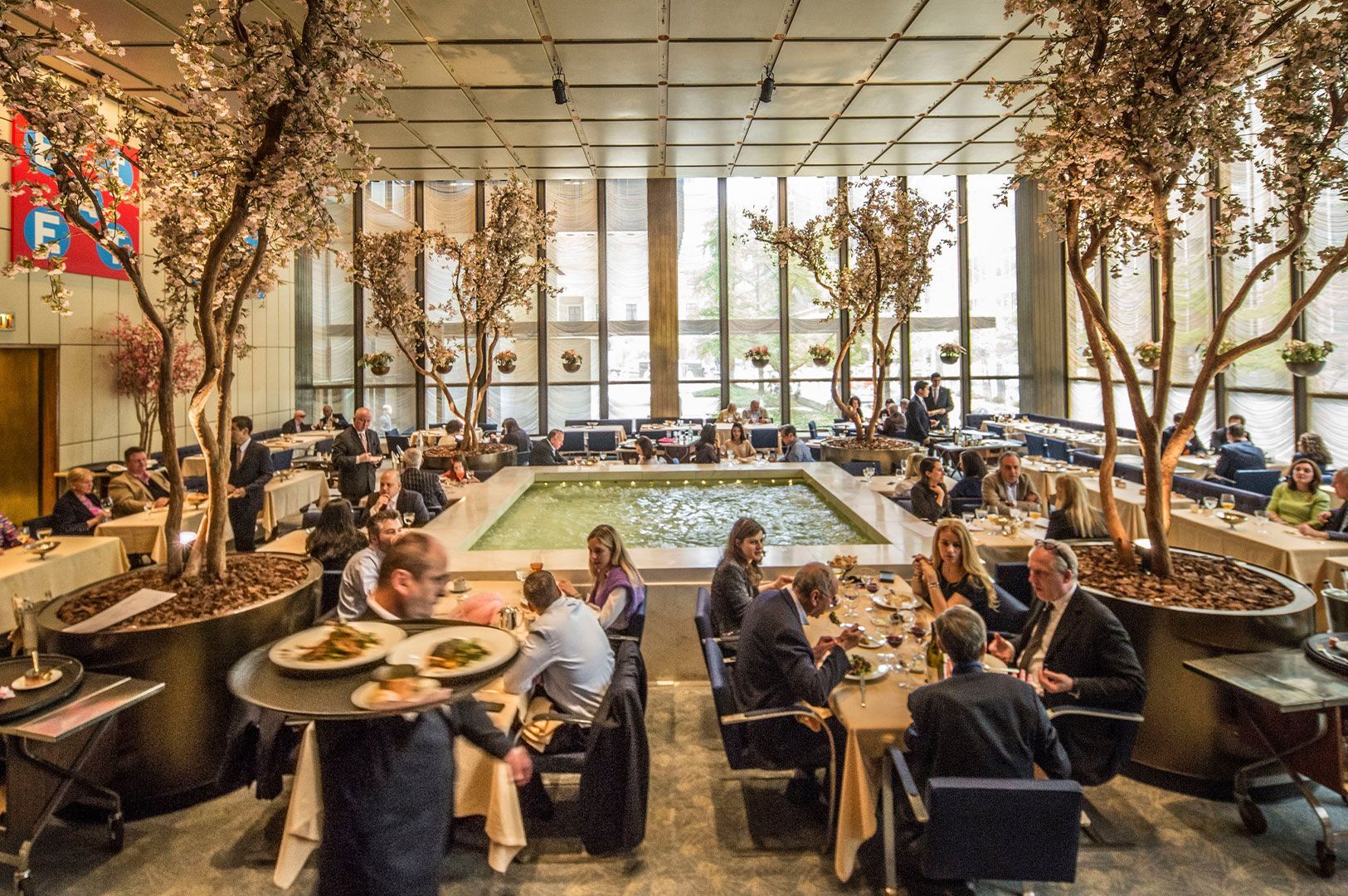 The iconic interior of The Four Seasons Restaurant Pool Room in New York, showing a large, rectangular water pool centerpiece surrounded by dining tables and mature trees in large planters.