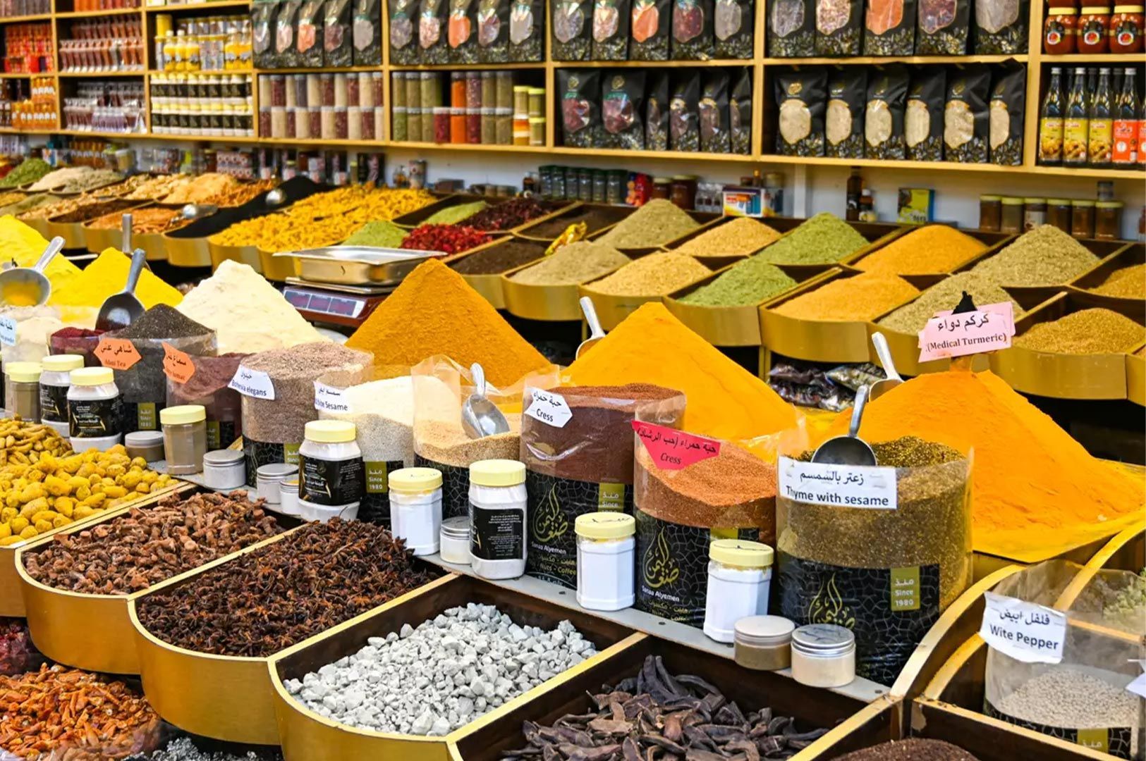 Vibrant piles of colorful spices including turmeric, thyme, and cress displayed in a traditional market with labeled jars in the background.