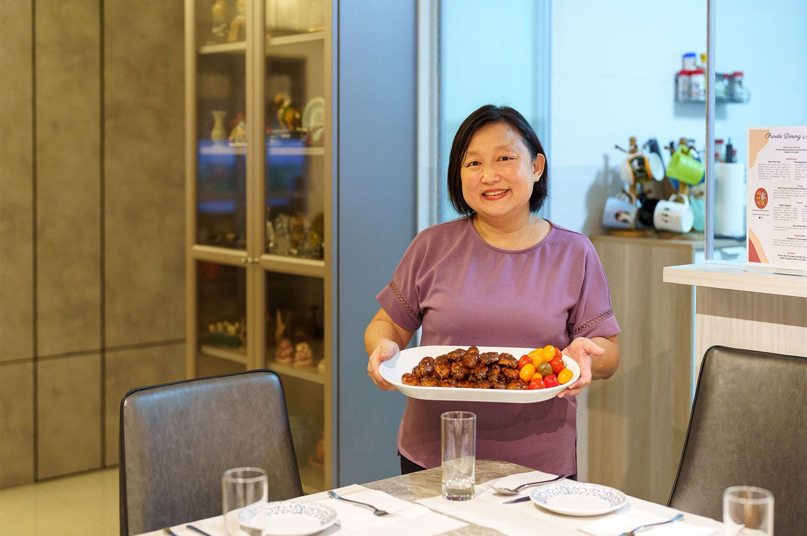 Smiling Asian woman standing at a dining table, holding and serving a white platter of dark, sticky glazed meat pieces (likely sweet and sour pork) garnished with cherry tomatoes.