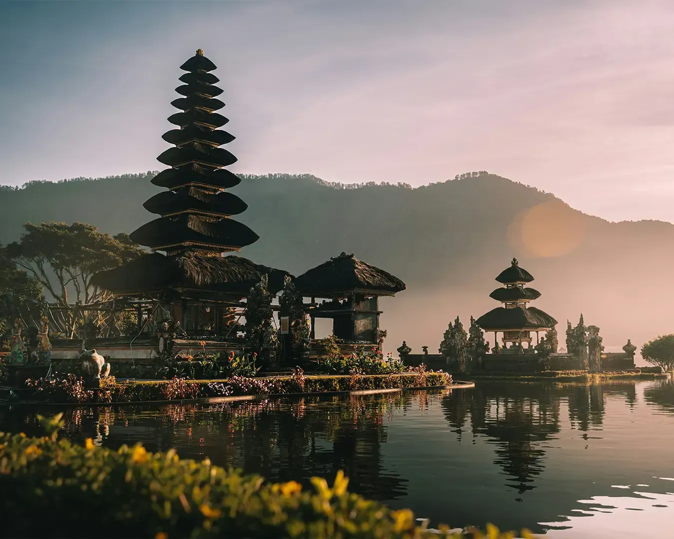 Text: Serene Balinese temple on water with tiered pagoda, silhouetted against misty mountains at sunset. Calm water reflects the peaceful scene.