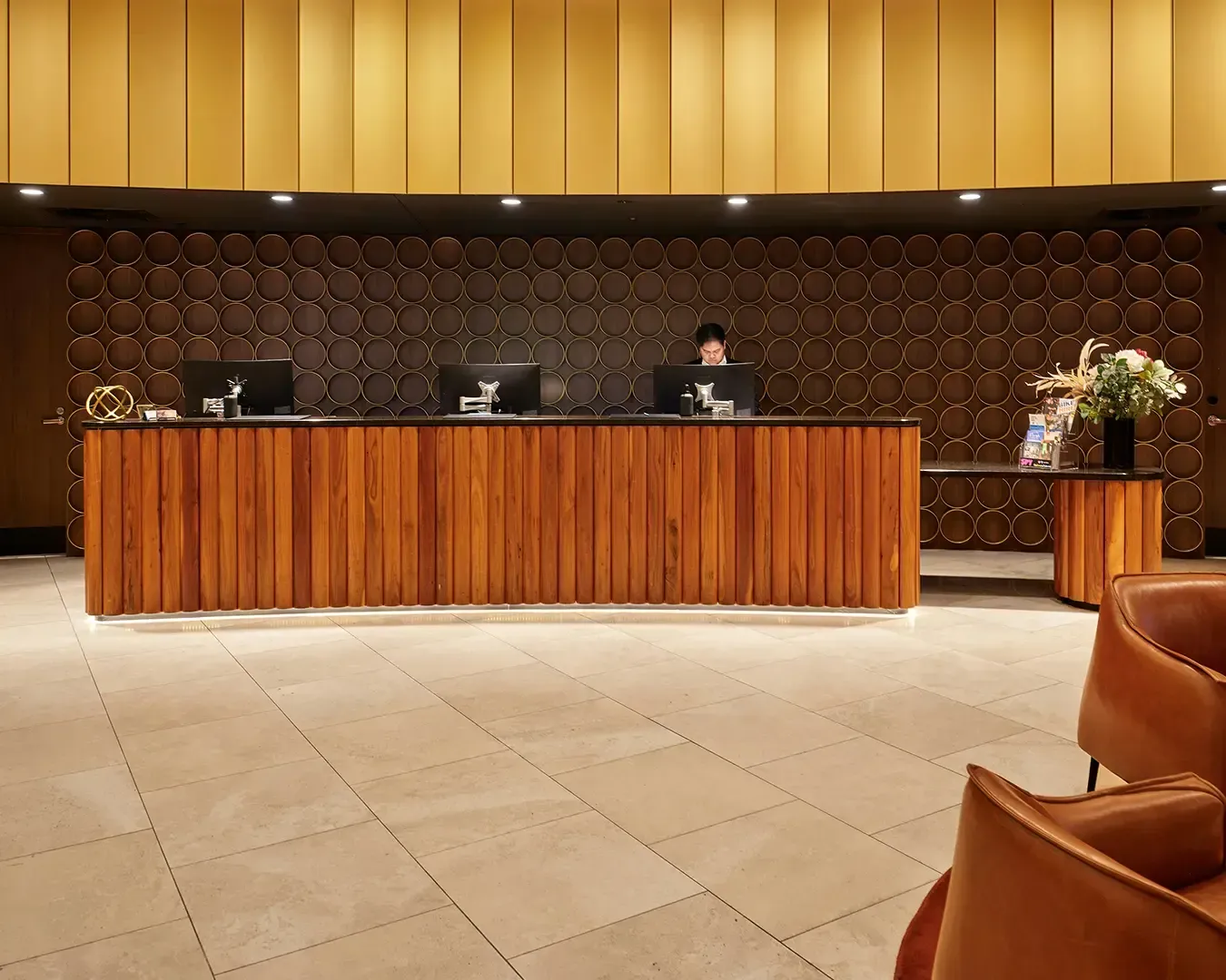 A modern hotel reception area with a warm ambiance. A wooden desk with computers and a receptionist is set against a circular-patterned wall.