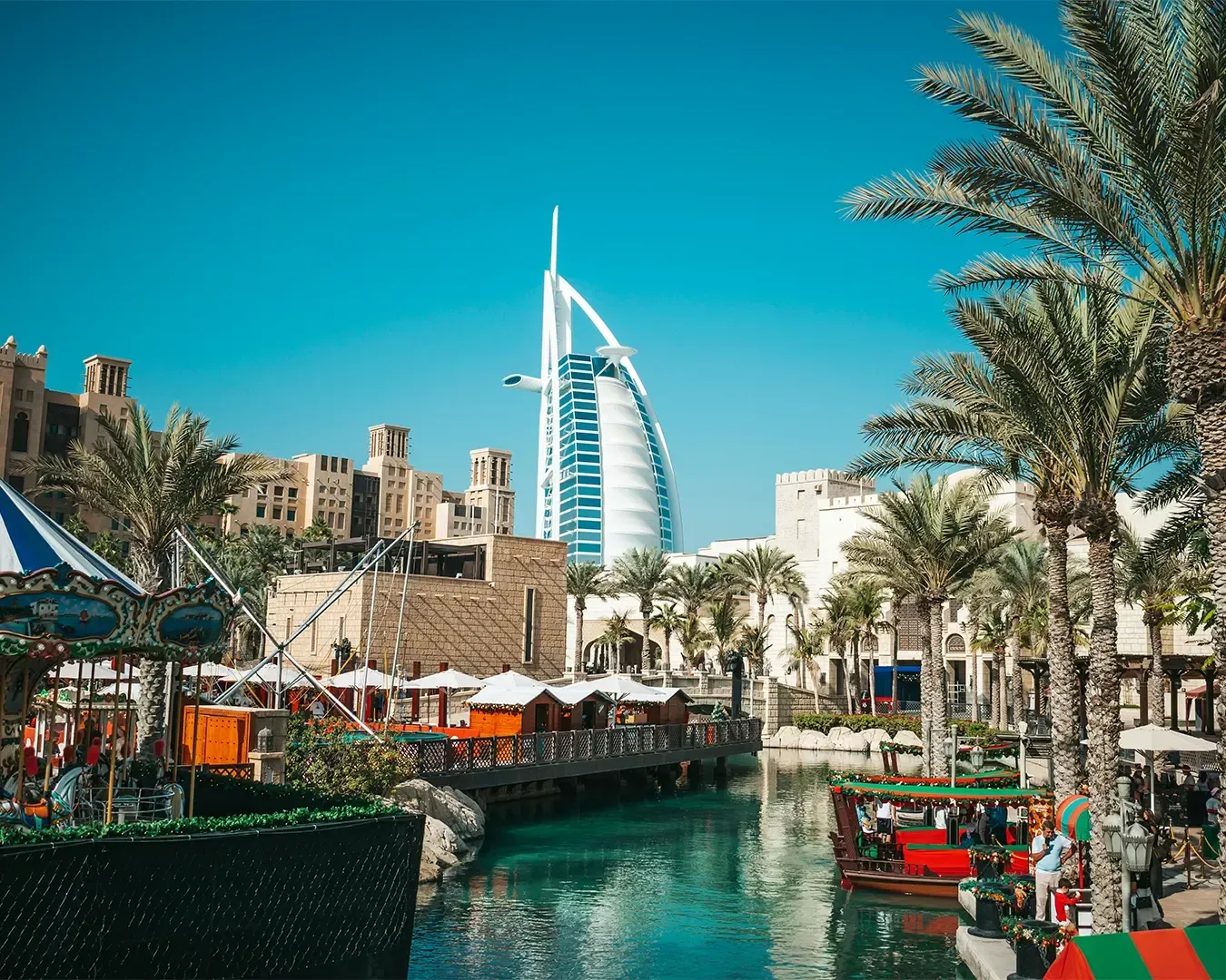 A bright day in Dubai features the iconic Burj Al Arab hotel in the background, with palm trees, traditional boats on teal water, and vibrant market stalls.