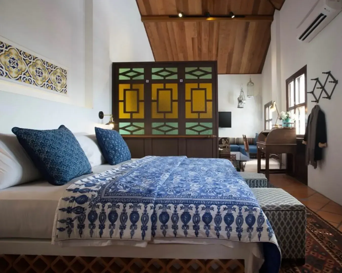 Cozy bedroom with a large bed adorned with blue patterned bedding, blue pillows, and a decorative screen. Warm lighting and a rustic wooden ceiling.