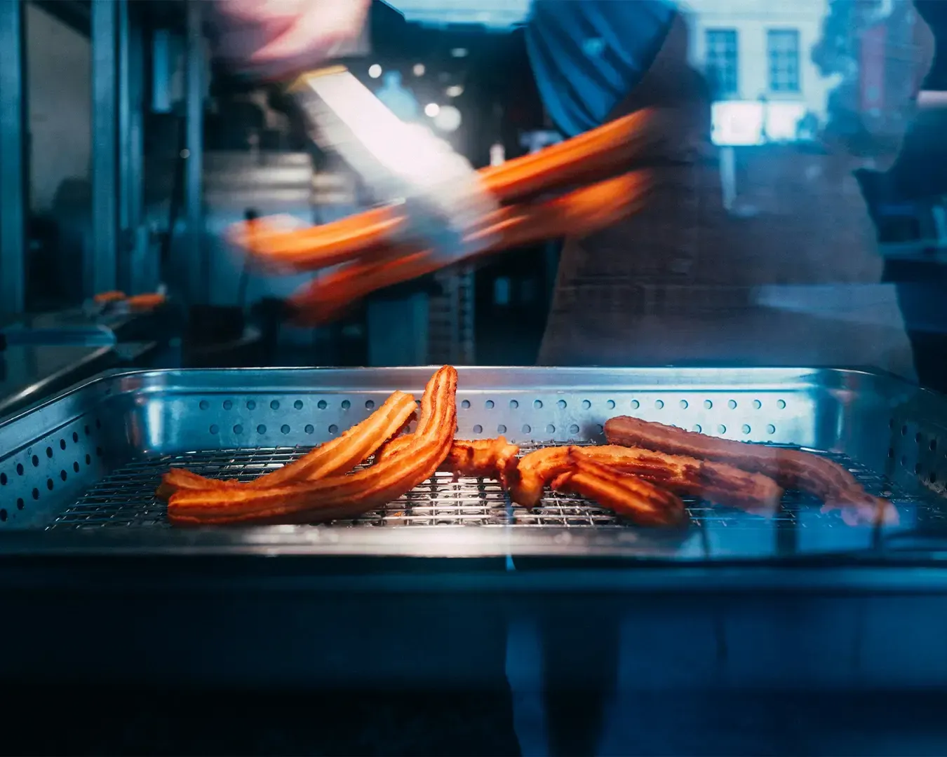 A street vendor preparing and selling freshly made churros, with golden pastries displayed or being fried. The scene feels warm and inviting, highlighting the crisp texture and sweet aroma of the snack in a casual street setting.