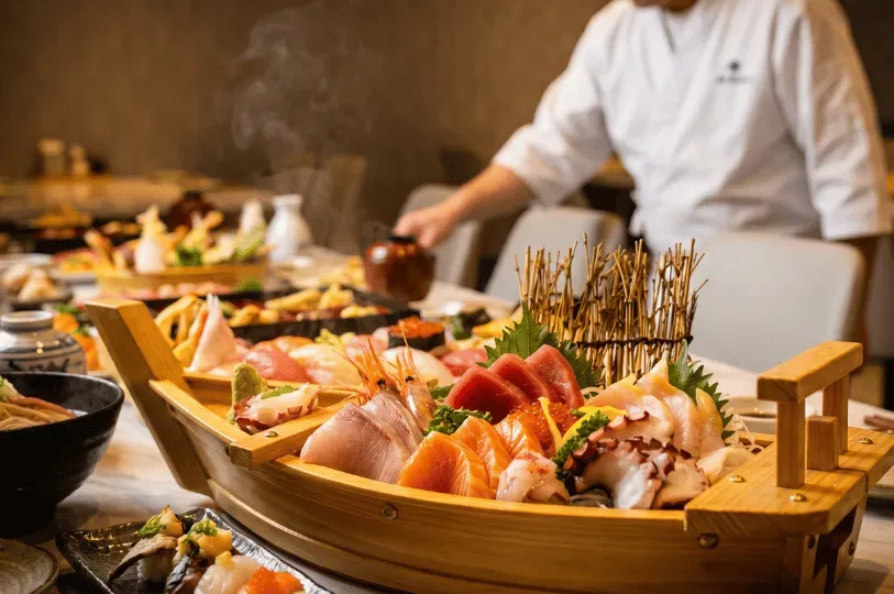 A magnificent wooden boat filled with fresh sashimi in sharp focus in the foreground, while a chef in a white uniform tends to the elaborate Chef's Table spread in the background.