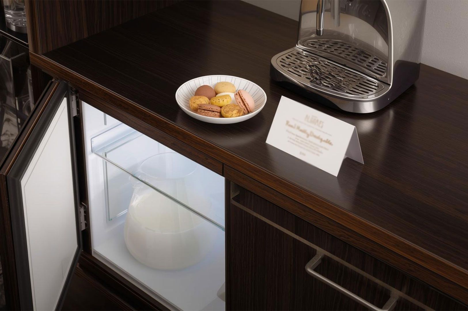 A hotel amenity station featuring a plate of colorful macarons, a welcome card, a chrome espresso machine, and an open mini-fridge.