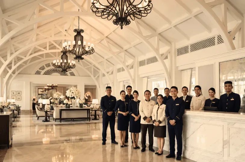 A large, coordinated team of over a dozen hotel staff members smiling warmly for the camera. They are standing behind and alongside a white marble reception desk in an elegant, airy lobby featuring high vaulted ceilings with white trusses and dark iron chandeliers.