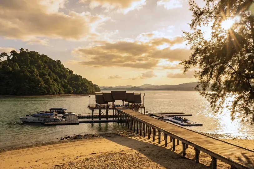 A long wooden pier extending from a sandy beach into calm ocean waters at sunset, featuring a traditional Thai-style pavilion at the end and several docked speedboats.