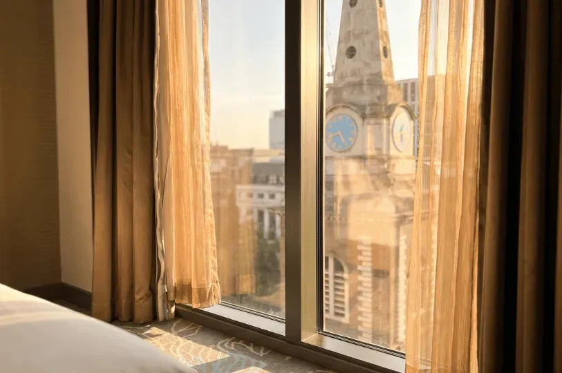 A view through a large hotel window framed by sheer golden curtains, looking directly across at a historic London stone church tower with a prominent blue-faced clock. Warm sunlight fills the room, casting patterns onto the carpeted floor below.
