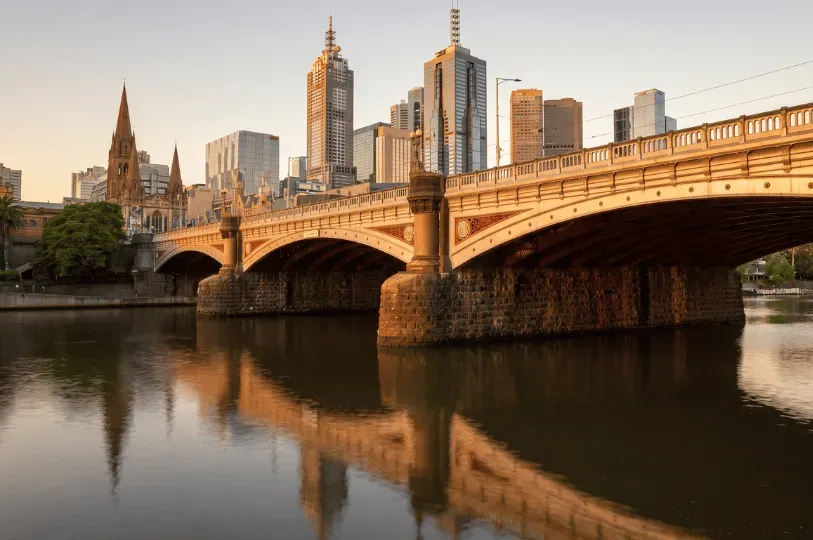 The Yarra River reflecting the stone arches of Princes Bridge and the Melbourne city skyline, including St Paul's Cathedral, at sunset.