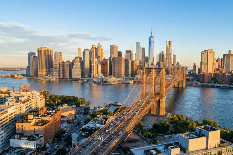 A sweeping aerial photograph of the Brooklyn Bridge spanning the East River, with the dense, modern skyline of Lower Manhattan illuminated by golden hour sunlight in the background.