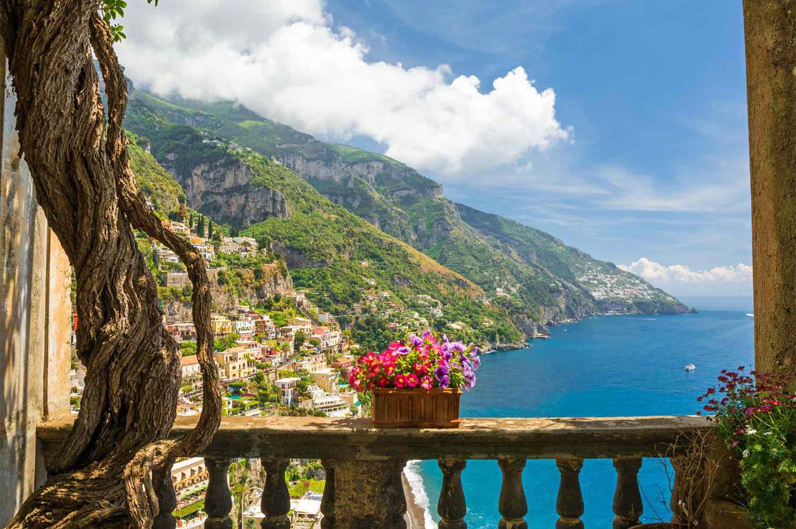 Scenic view of Positano on the Amalfi Coast from a shaded balcony, showing the cliffside houses, the blue sea, and a twisted vine framing the scene.