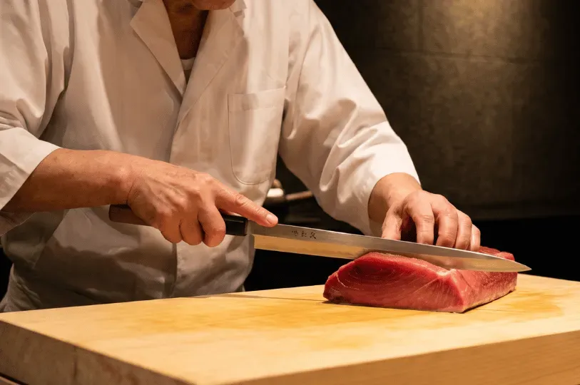 A close-up of a sushi chef using a long, slender, and highly polished yanagiba knife to slice through a large, pristine loin of ruby-red tuna on a wooden cutting board, capturing the precision and skill of professional Japanese knife work.
