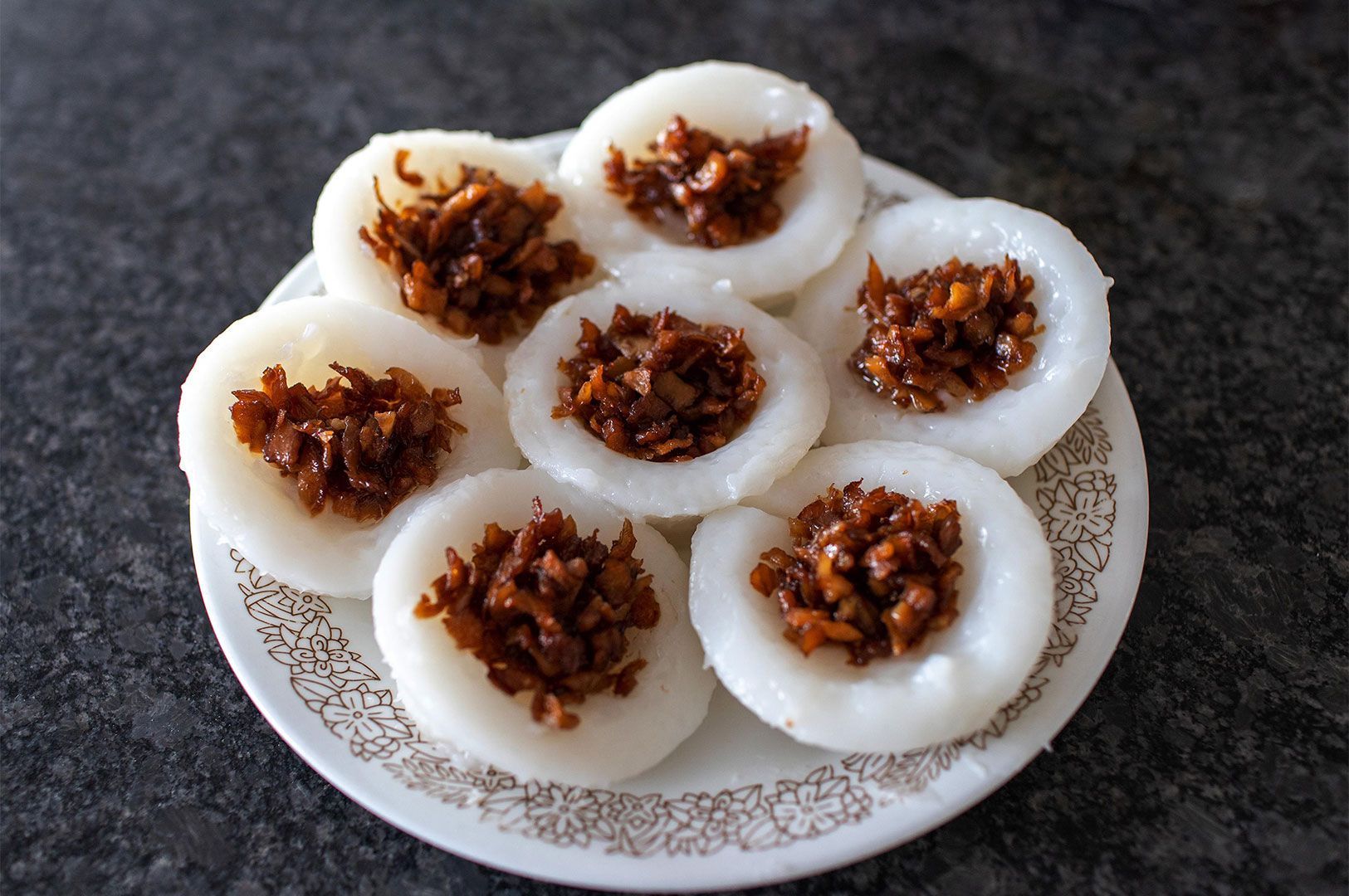 A small white plate holding seven Vietnamese Bánh Bột Lọc (savory steamed rice cakes/dumplings) topped with a dark, savory mixture of caramelized shrimp and/or pork, on a dark countertop.