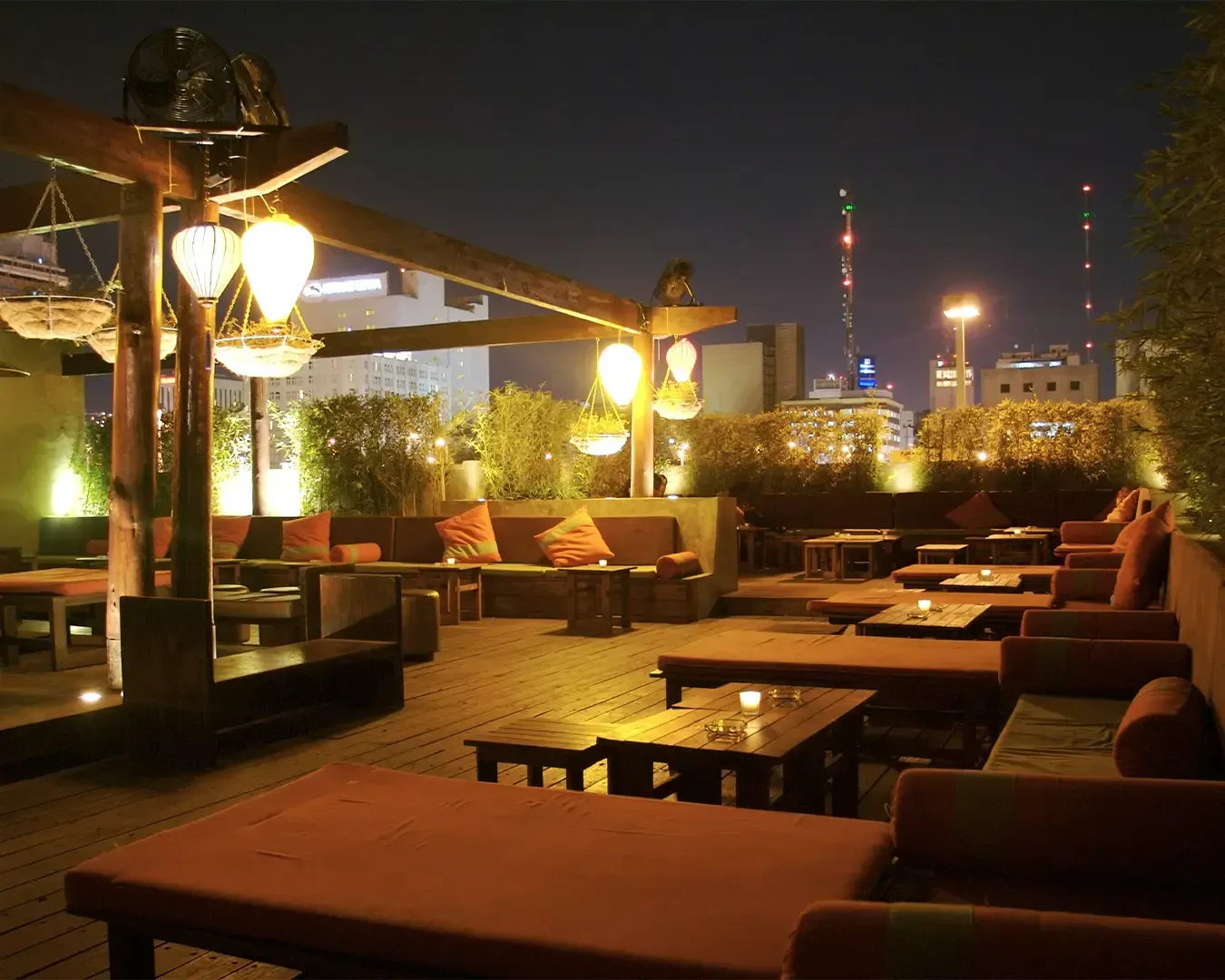 Chic rooftop lounge at night with cozy seating, warm lighting, and cityscape views. Ambient atmosphere with glowing lanterns and soft cushions.