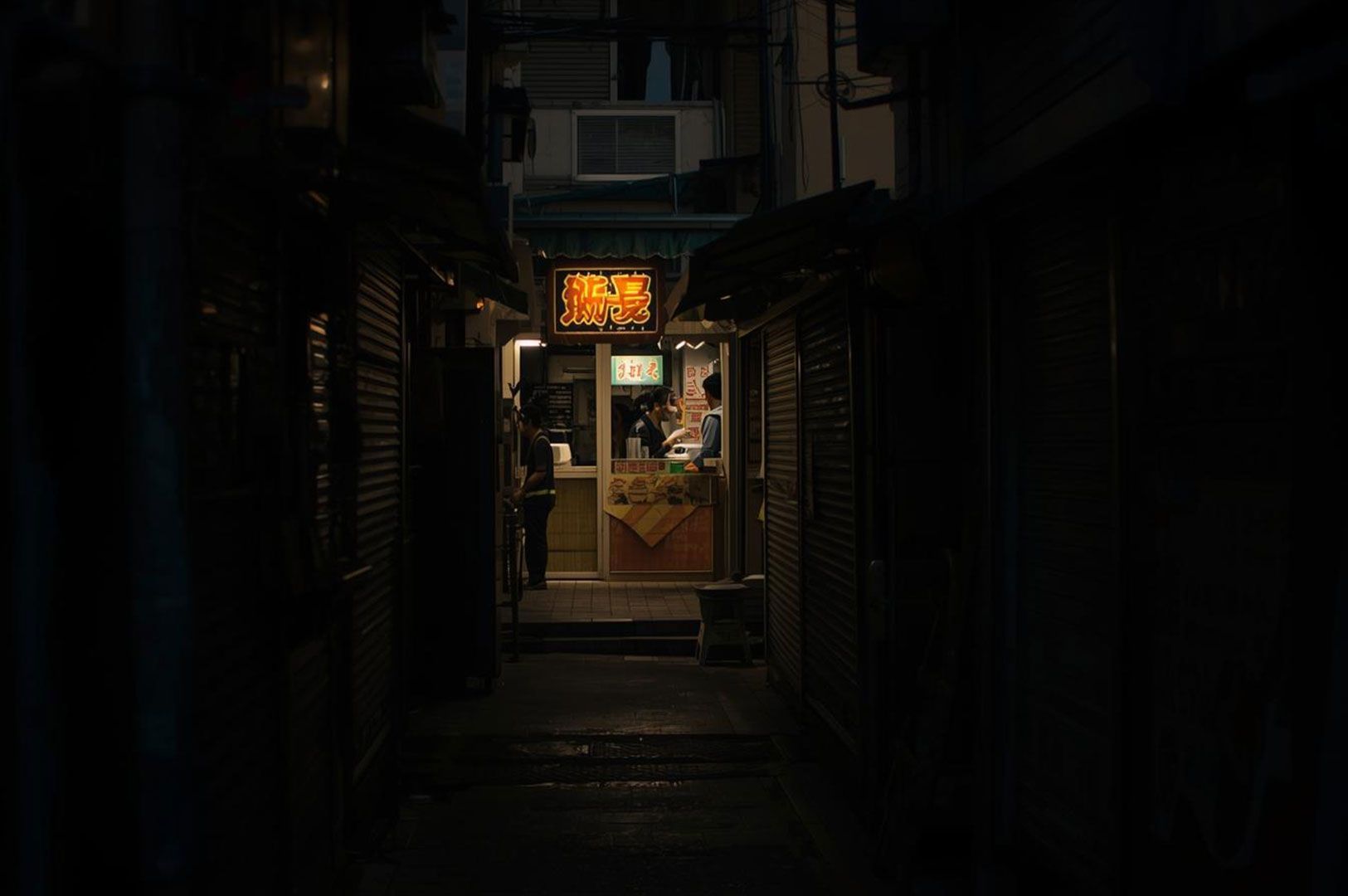 A dark, moody shot looking down a narrow, shadowy city alley, with a brightly lit street food stall and a Chinese neon sign glowing at the end of the passage.
