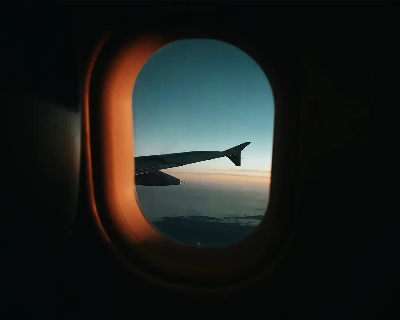 A view from inside an airplane looking out through the window at the aircraft wing extending into the sky. Clouds and open sky create a peaceful in-flight perspective.