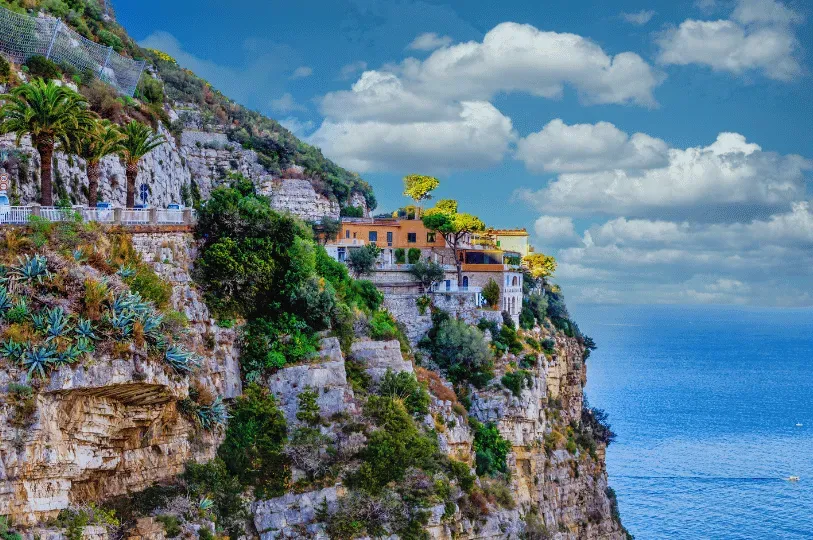 A terracotta-colored luxury villa built into the steep, lushly vegetated cliffs of the Amalfi Coast, overlooking the ocean under a cloudy blue sky.